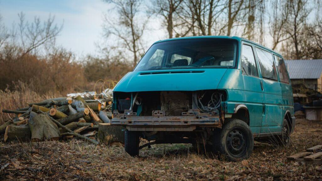 Une vieille camionnette abandonnée se trouve dans une zone boisée avec des bûches coupées, évoquant une apparence rustique et négligée.